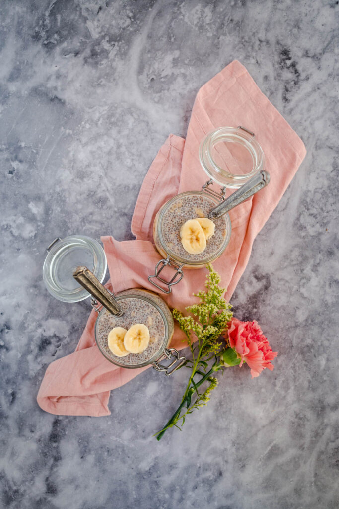Two jars of chia pudding garnished with banana slices on a marble surface with a pink cloth and a small pink flower to the side.