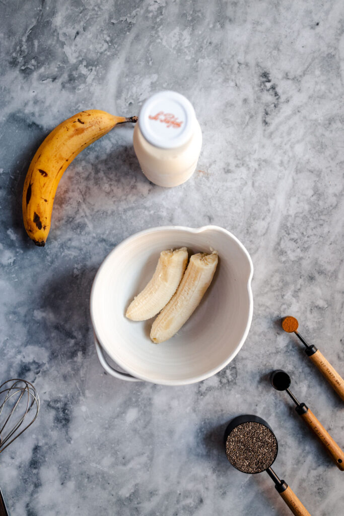 A banana, a bottle of milk, a bowl with banana slices, and baking utensils on a marble countertop.