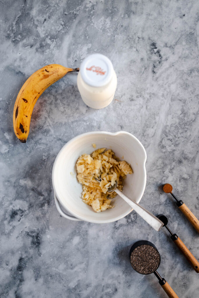 Overhead view of a bowl with mashed bananas, a whole banana, a bottle of milk, and a coffee grinder on a marble surface.