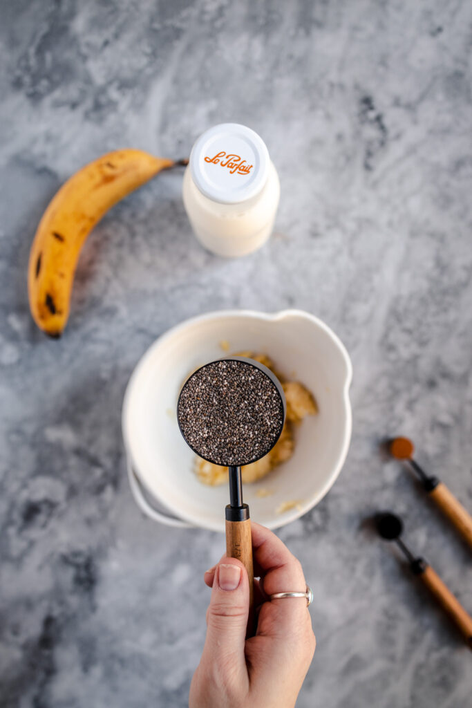 A person holding a measuring spoon with chia seeds over a bowl next to a ripe banana and a jar of yogurt.