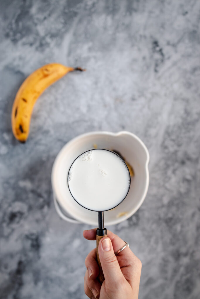 Hand frothing milk in a white container with a ripe banana in the background on a gray surface.