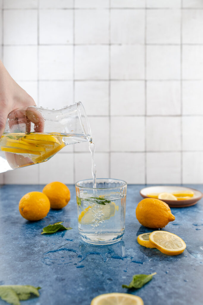 Pouring lemon-infused water into a glass on a kitchen counter with whole and sliced lemons and mint leaves around.