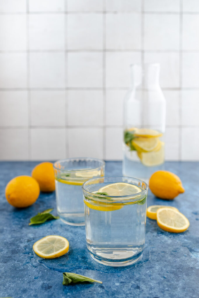Two glasses of lemon water with fresh lemons on a blue countertop.