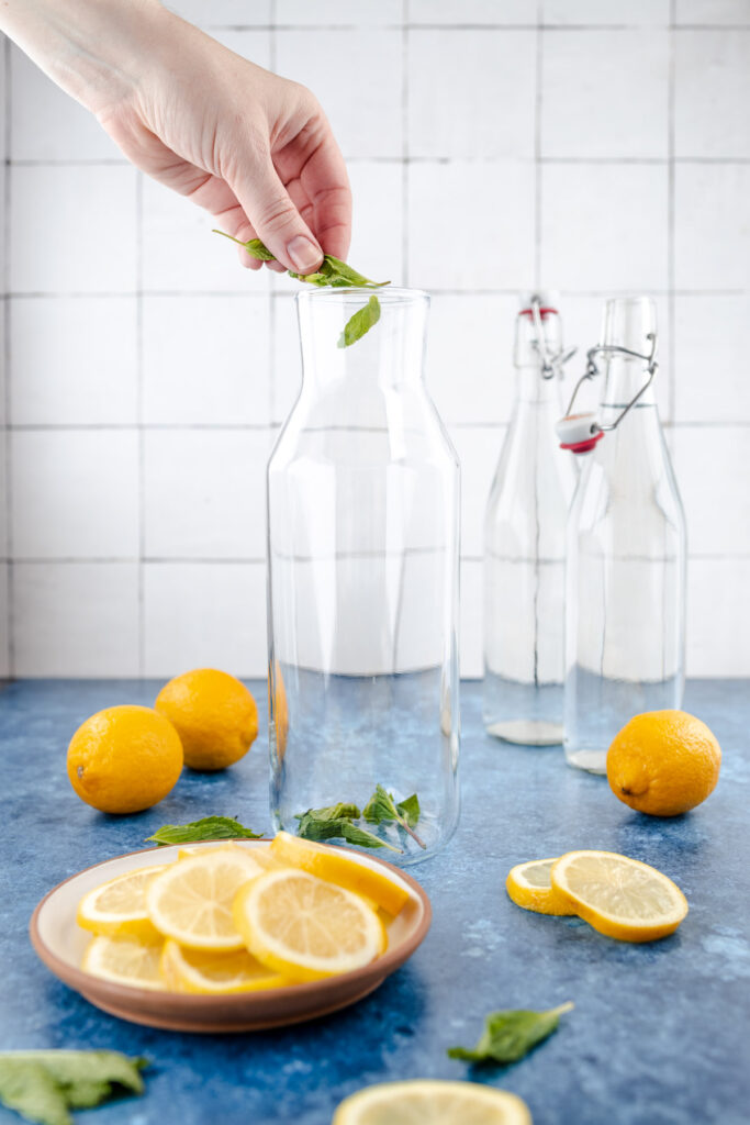 A person adding a mint leaf to a bottle of water, with sliced lemons and basil leaves on a tabletop.