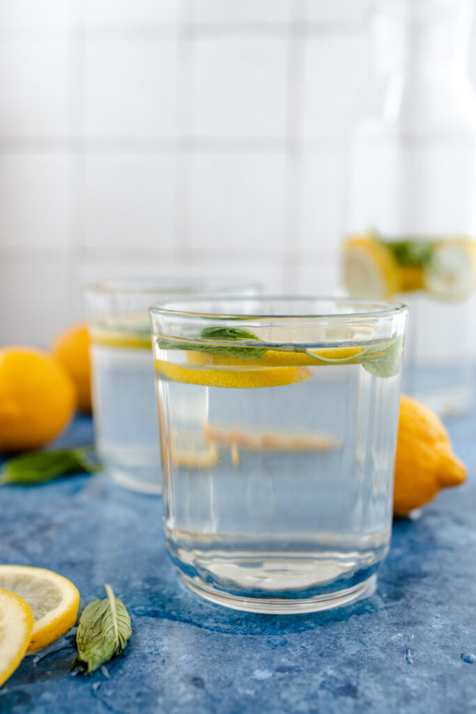 Two glasses of water with lemon slices and mint on a blue surface with more lemons in the background.