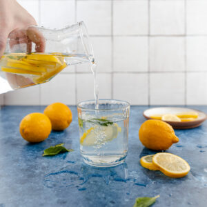 Pouring lemon water into a glass with mint on a blue surface surrounded by fresh lemons.