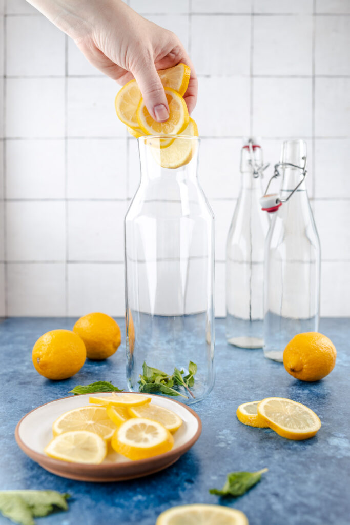 A person placing lemon slices into a glass bottle, with fresh lemons and mint leaves on the side.