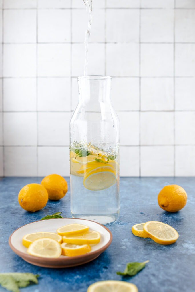 A carafe being filled with water, with slices of lemon and mint leaves inside, beside a plate of lemon slices and whole lemons on a blue surface with a tiled background.