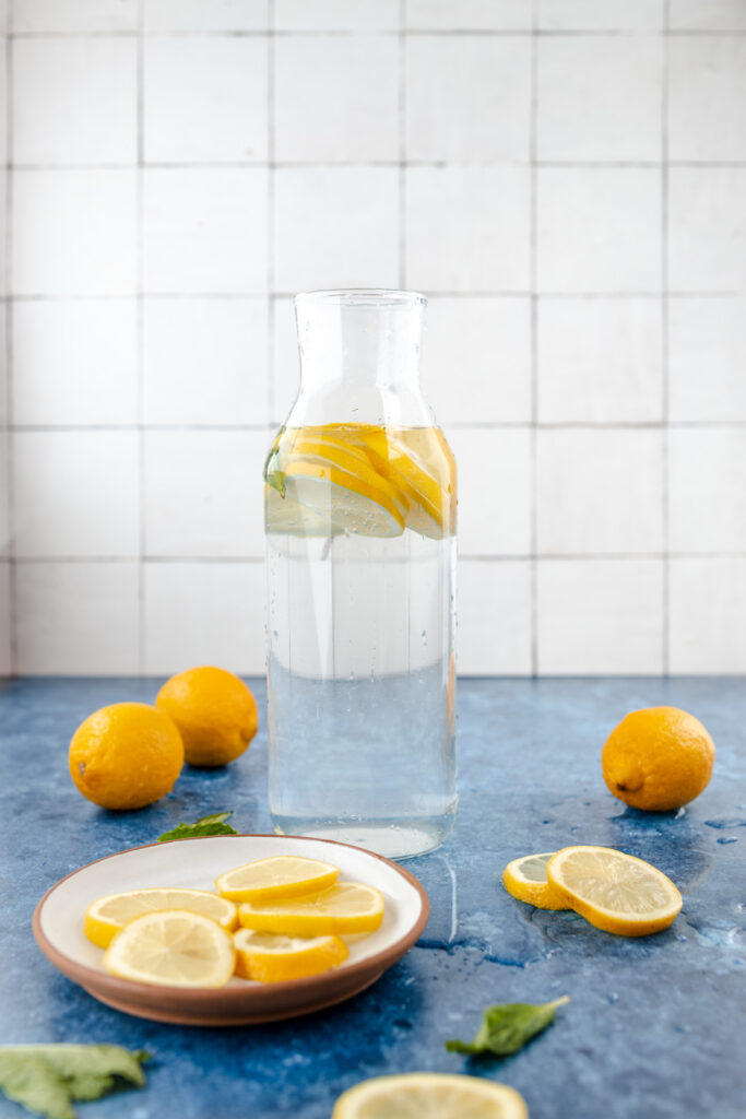 Glass bottle of water infused with lemon slices and mint leaves, accompanied by a plate of lemon slices, whole lemons, and scattered mint leaves on a blue surface with a tiled backdrop.