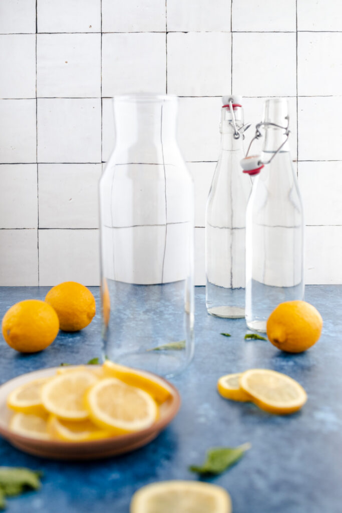 Clear glass carafe and bottle with water accompanied by fresh lemons on a blue surface against a white tiled backdrop.