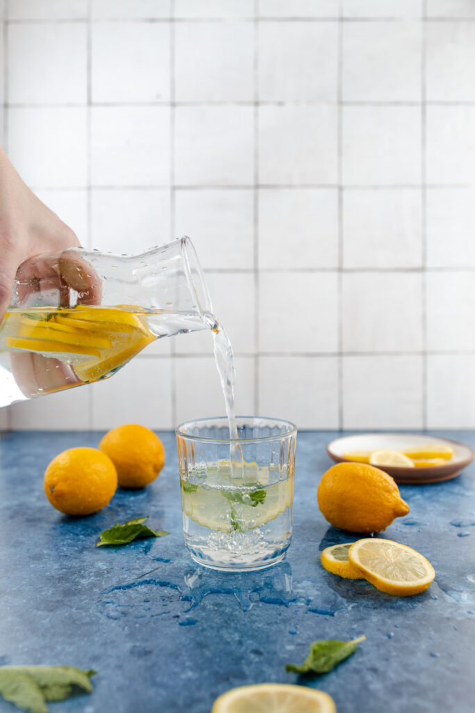 Pouring lemon-infused water into a glass on a kitchen counter with fresh lemons and mint leaves around.