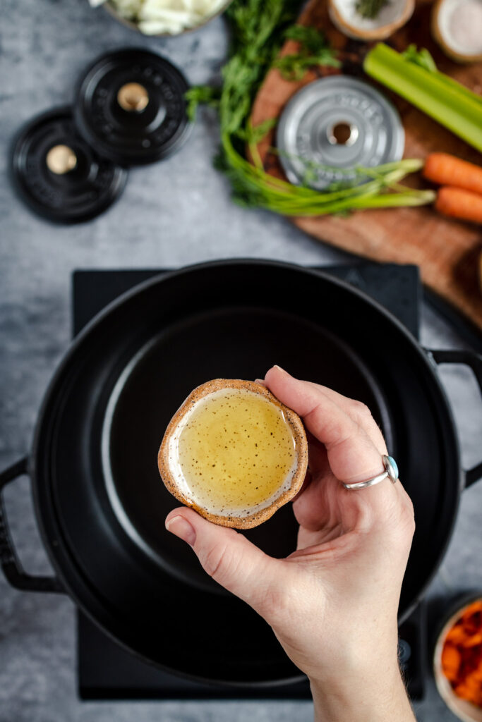 A hand holding a small bowl of broth above a stove with ingredients and cookware in the background.