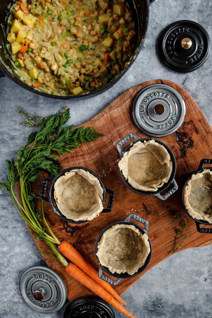 Preparation of individual pot pies with filling in a pot and pie crusts in small ramekins, alongside fresh carrots and herbs on a cutting board.