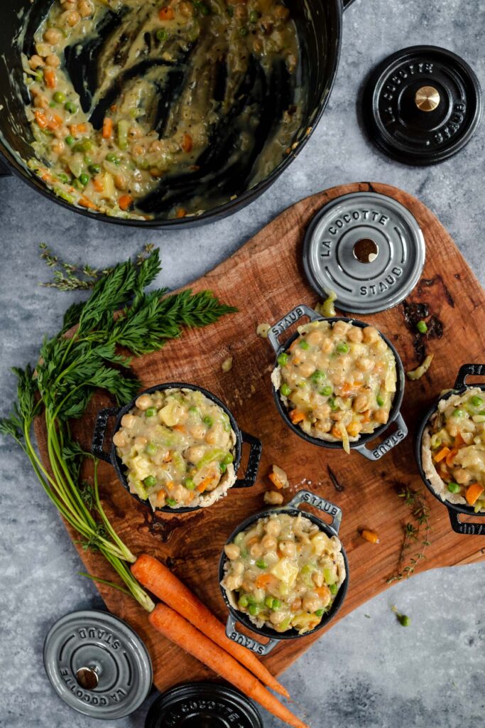 Mini pot pies with vegetable filling on a wooden board next to fresh herbs and ingredients, with a mixing bowl in the background.