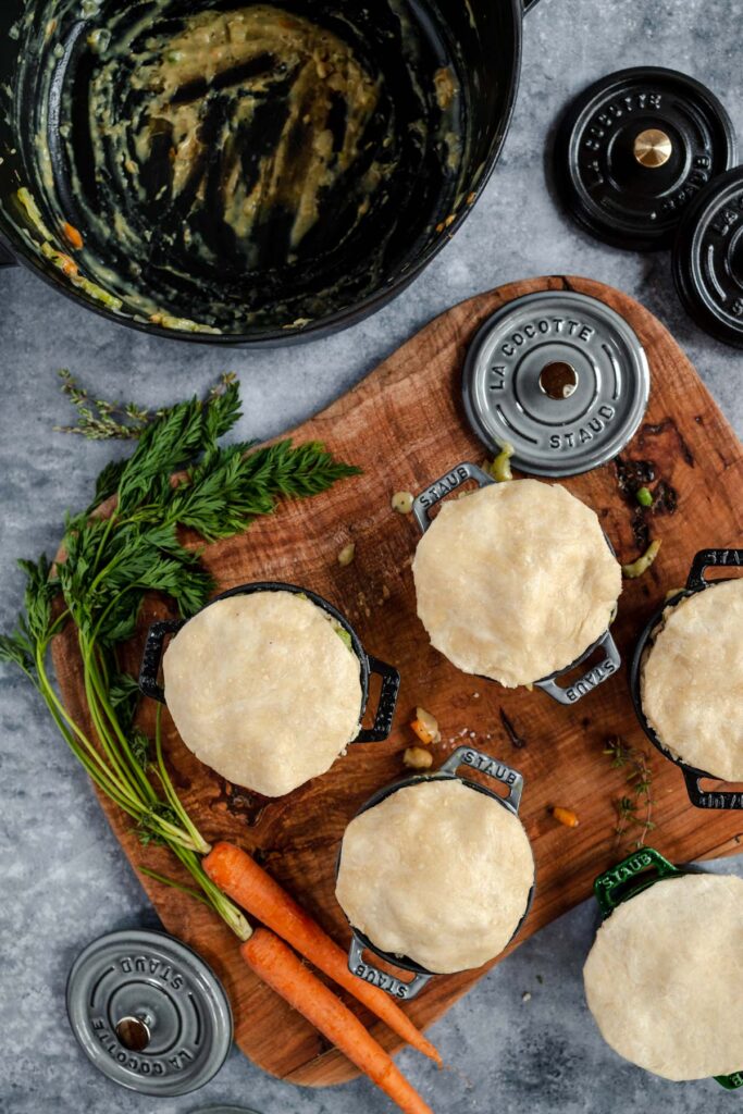 Unbaked pies placed in metal molds on a cutting board alongside carrots and fresh herbs, with an empty mixing bowl and lids nearby.