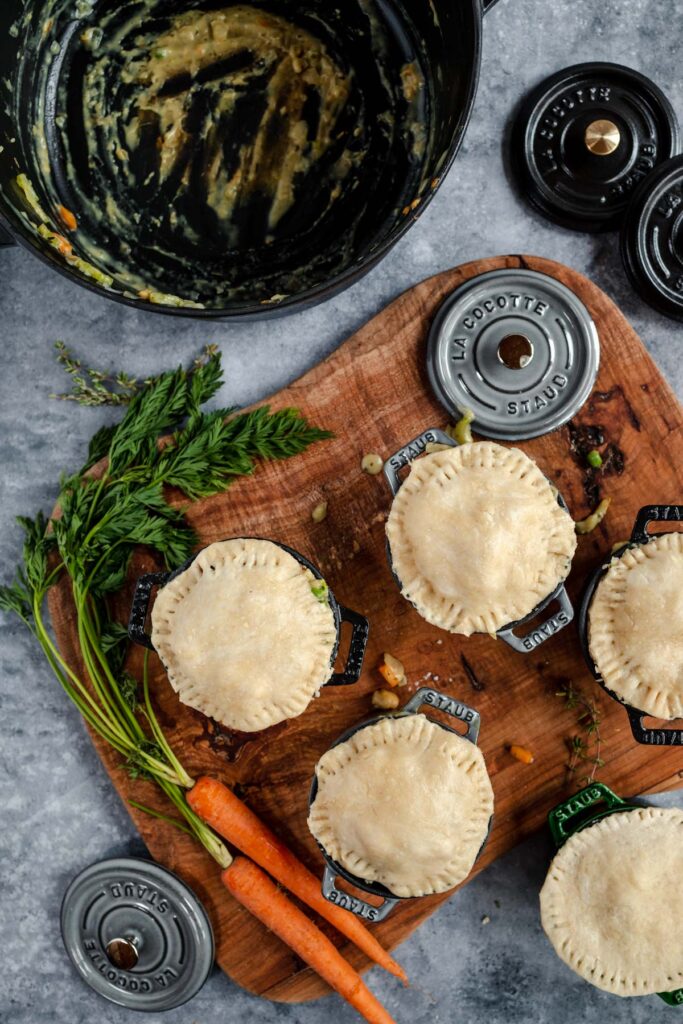 Freshly prepared uncooked pies on a wooden board with carrots and herbs, next to an empty bowl and cooking weights.