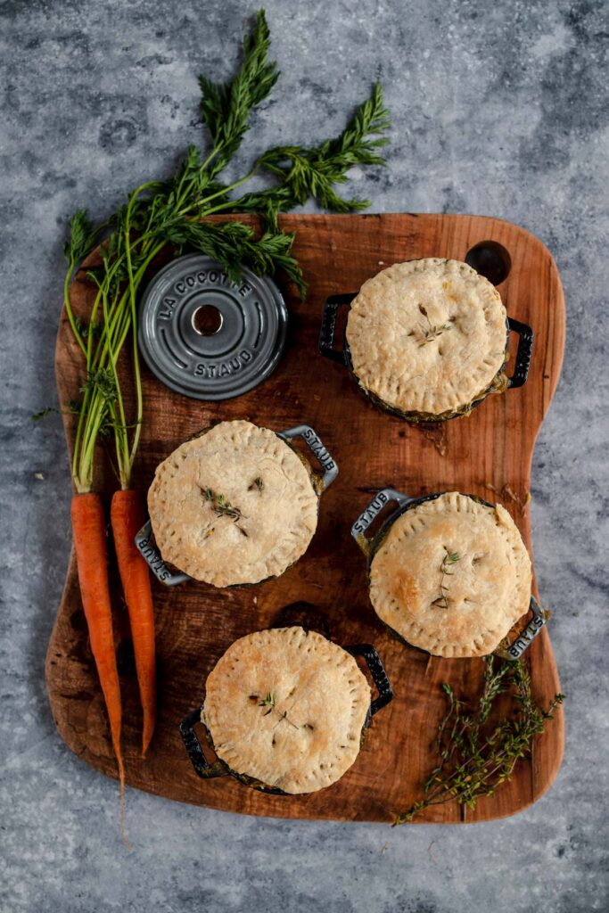 Four savory pies on a wooden board with carrots and herbs.