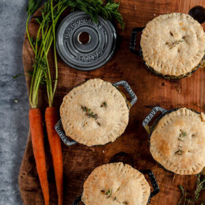 Four mini pot pies with a sprig of thyme on a wooden cutting board, accompanied by fresh carrots and dill, with a staub cast iron pot lid in the background.