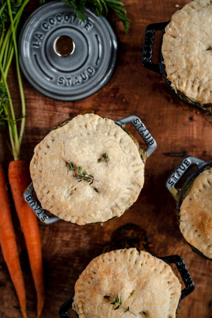 Individual pot pies in black ramekins garnished with thyme, accompanied by fresh carrots and a sprig of herbs on a wooden surface.