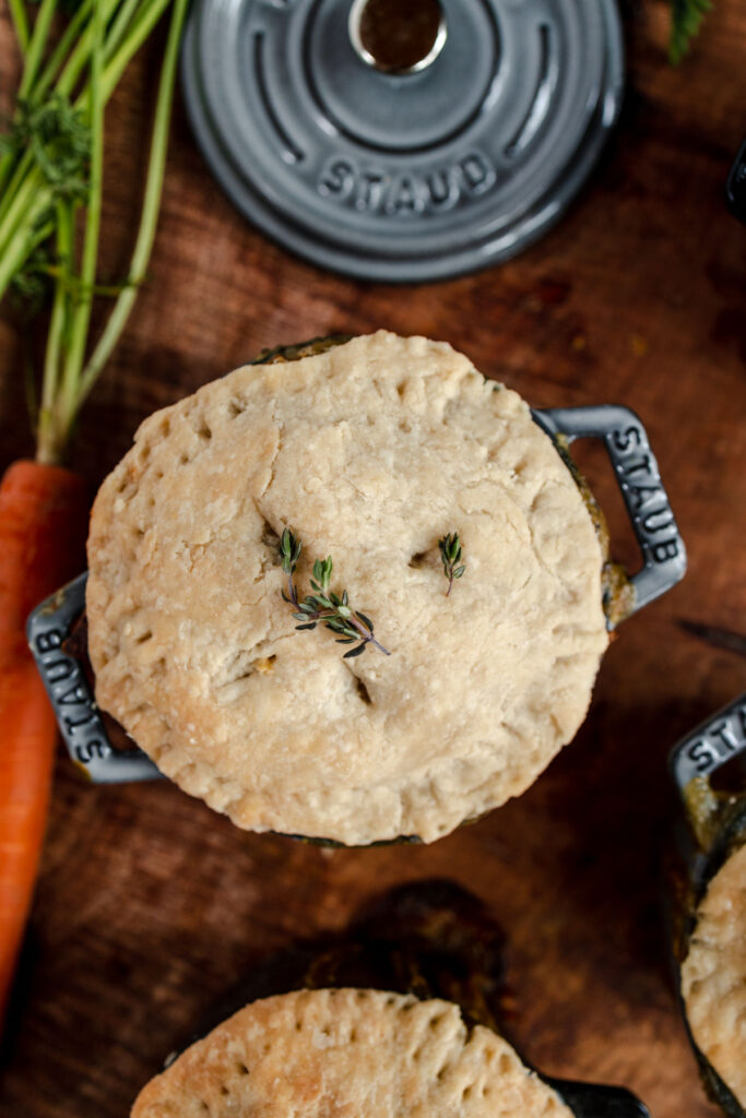 Top view of a pot pie in a small cast-iron skillet garnished with thyme, surrounded by fresh carrots and herbs on a wooden surface.