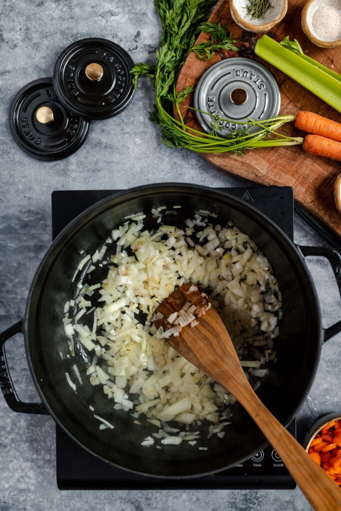 Sautéing diced onions in a black pan with a wooden spoon, with carrots, celery, and herbs nearby.