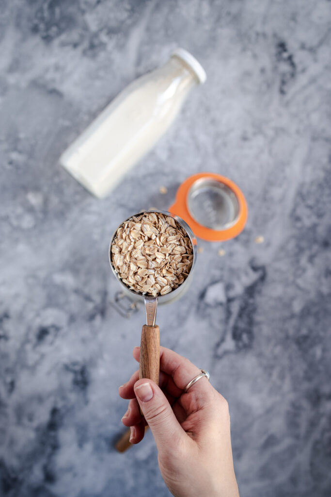 A hand holding a measuring cup filled with rolled oats over a marble surface, with a bottle of milk and a lid in the background.