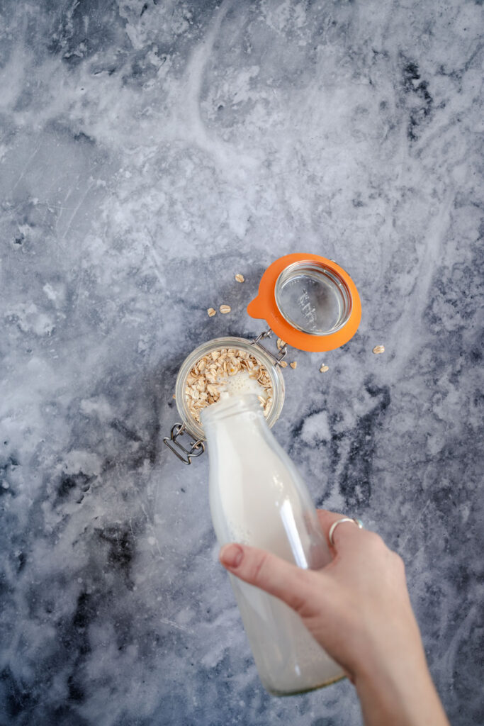 A person holding a bottle of milk next to a glass jar of oats and an orange sifter on a marble surface.