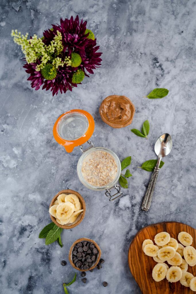 Overhead shot of a breakfast setup with oatmeal, banana slices, peanut butter, and chocolate chips on a marble surface, complemented by fresh flowers.