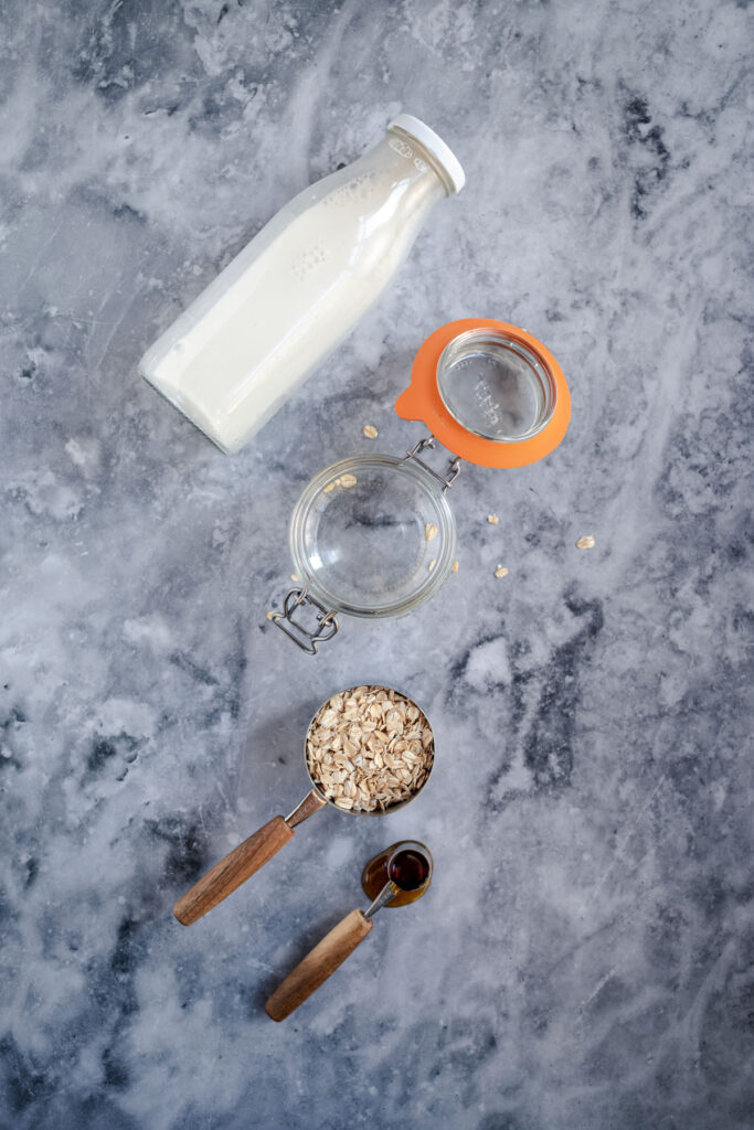 A bottle of milk, an open empty jar, a scoop of oats, and a spoonful of honey on a marble surface.