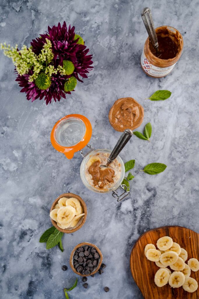An overhead shot of a food preparation scene with ingredients for a sweet recipe, featuring bananas, chocolate chips, peanut butter, and honey, arranged neatly on a marbled surface next to a bouquet of flowers.