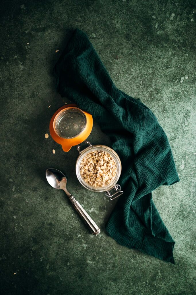 Overhead view of a breakfast setup with a jar of granola, a glass of orange juice, a spoon, and a green napkin on a textured surface.