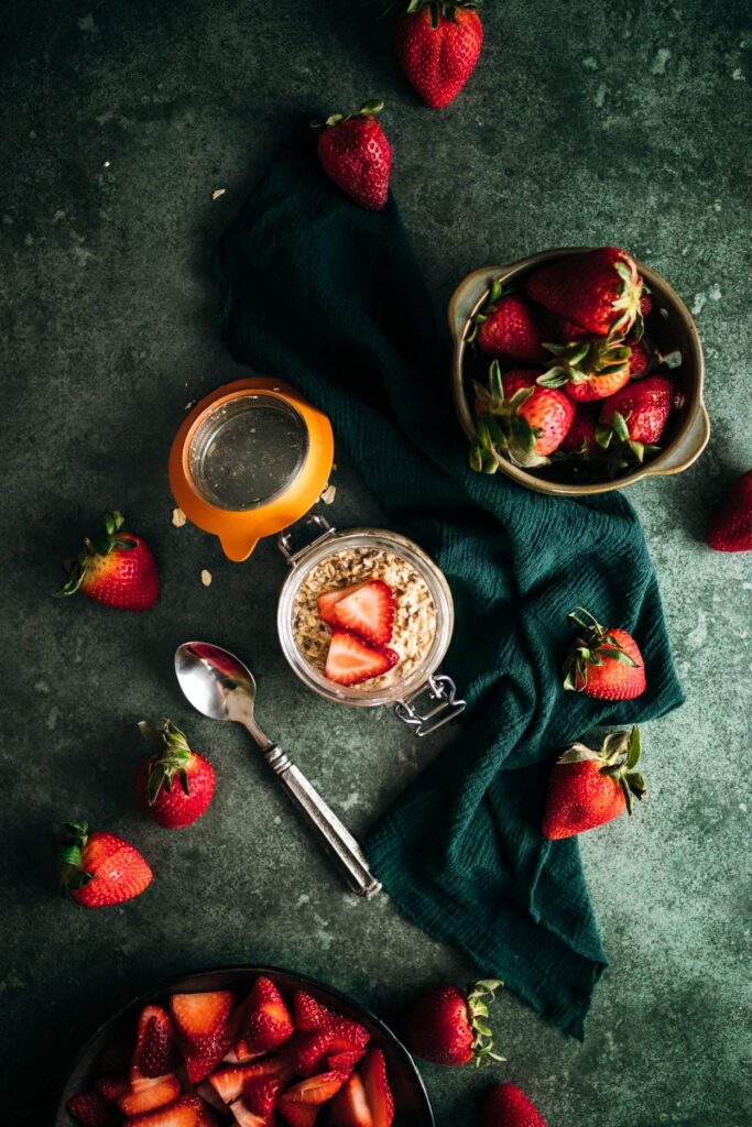 A flat-lay image of fresh strawberries on a textured surface with a jar of overnight oats and a green cloth napkin.