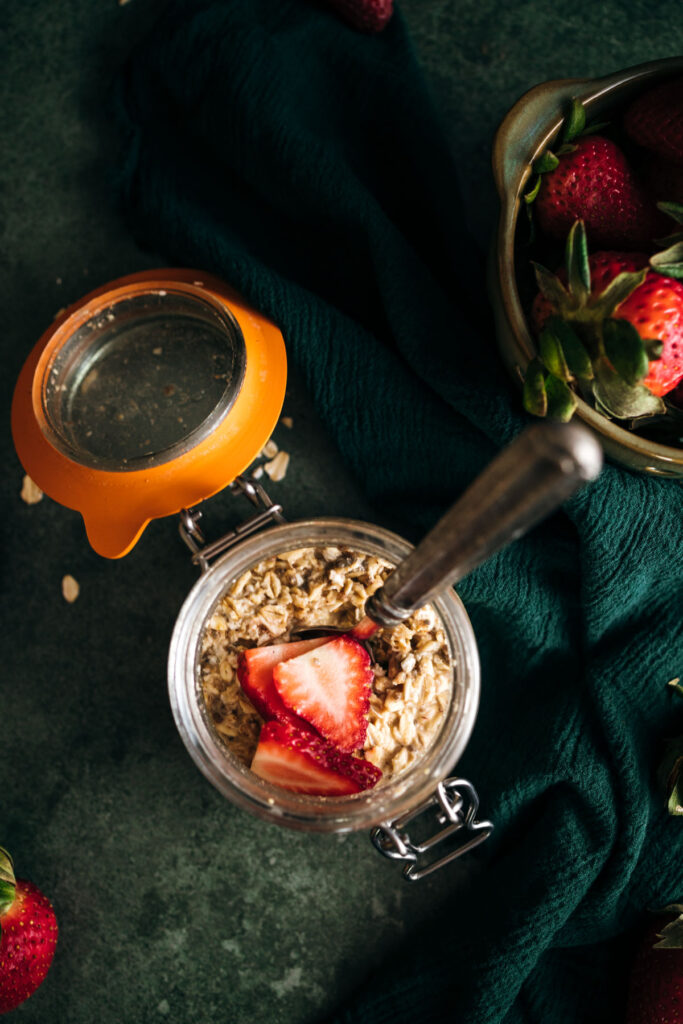 Overhead view of a jar of granola with fresh strawberry slices on a dark green cloth, surrounded by whole strawberries.