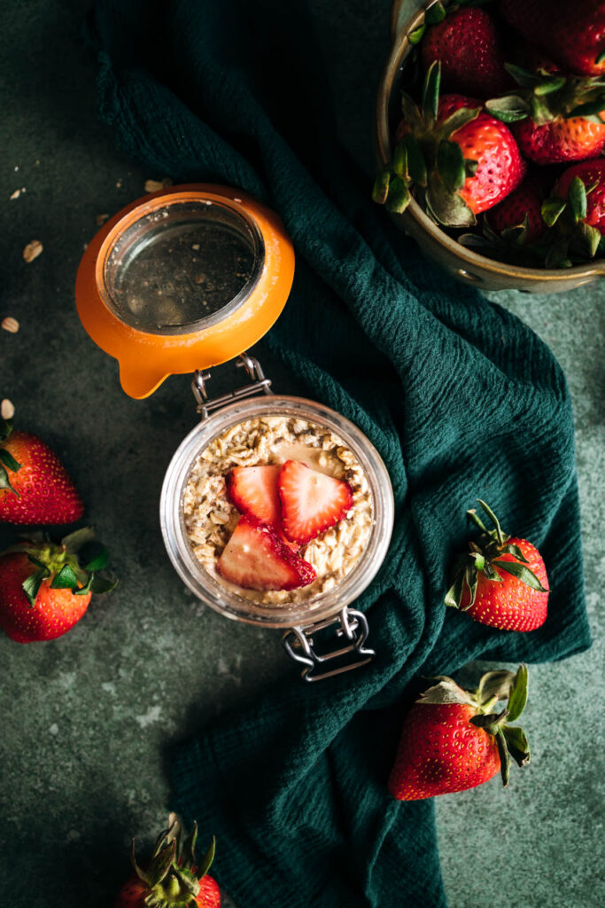 A glass jar of oatmeal topped with sliced strawberries, surrounded by fresh strawberries and a blue cloth.