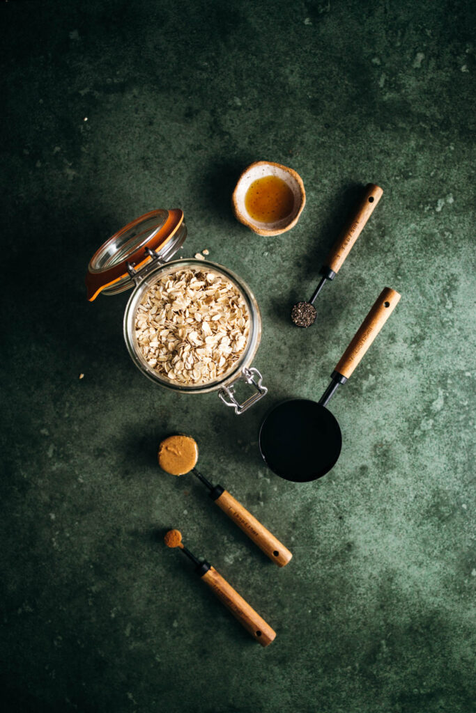 Ingredients and utensils laid out for preparing a recipe, featuring oats, spices, and a measuring cup on a green textured surface.