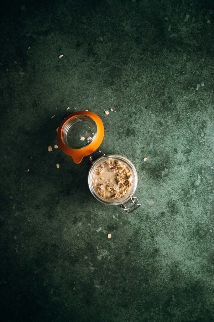An open glass jar filled with granola on a textured green background, with some granola spilled on the surface.