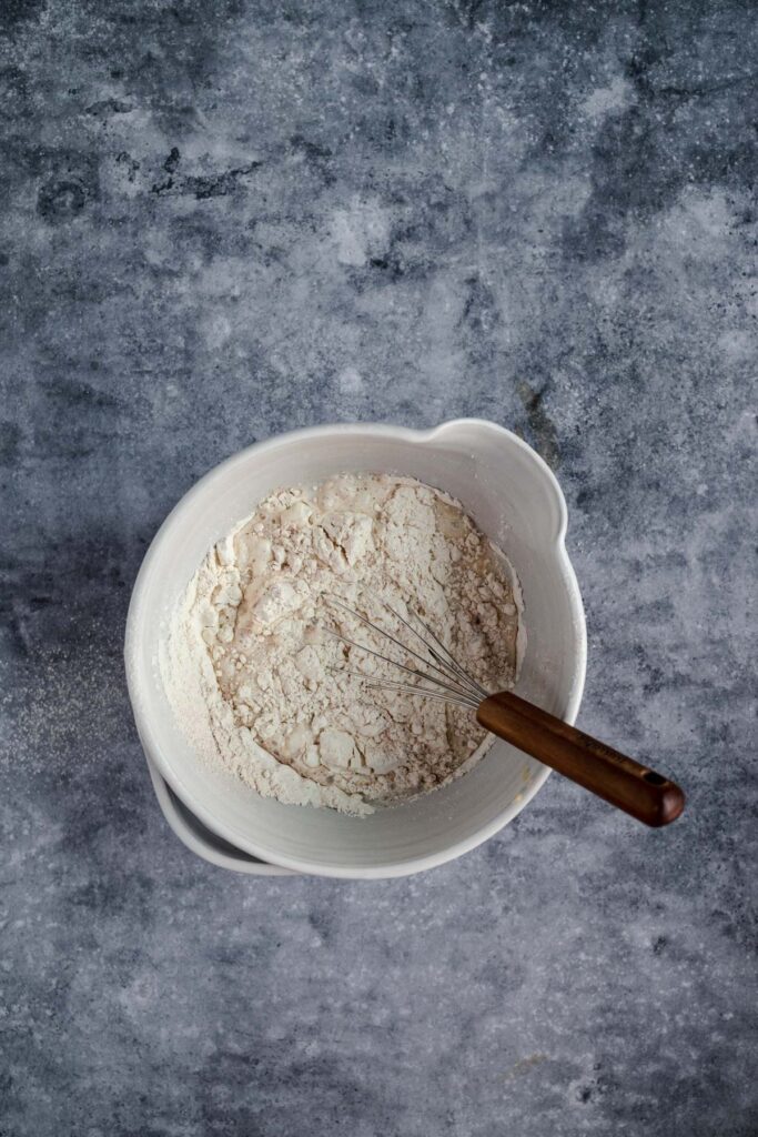 A bowl of flour with a metal whisk on a textured gray surface.