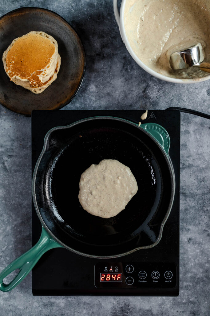 A pancake being cooked on a cast iron skillet over an induction stove with a bowl of batter and a plate with a cooked pancake nearby.