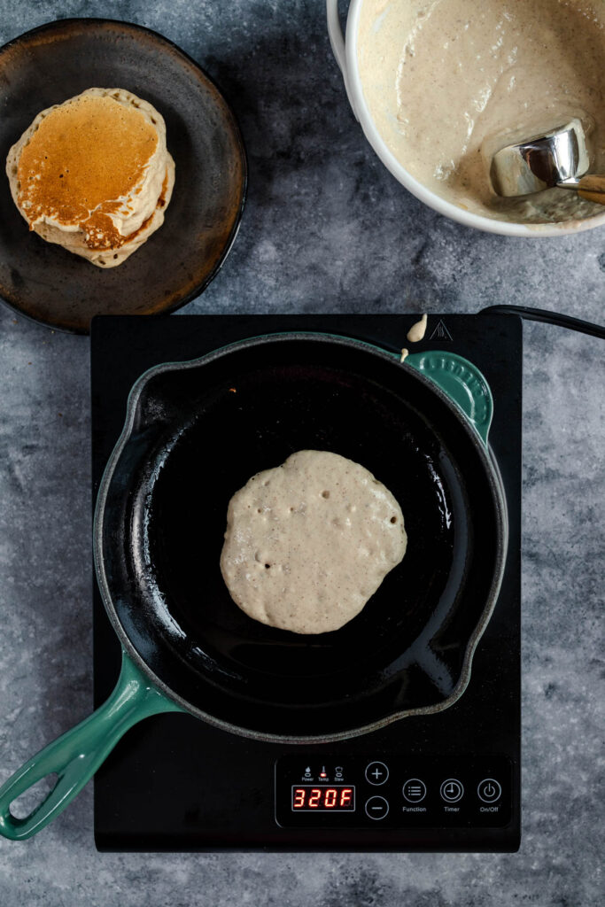 A pancake being cooked on a portable induction cooktop with a bowl of batter and a cooked pancake on a plate in the background.