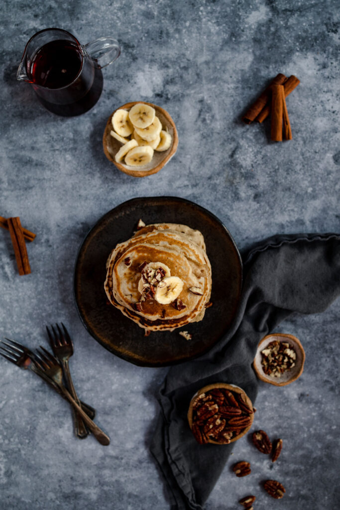 Stack of pancakes with banana slices and nuts on a rustic table setup with syrup and cinnamon sticks.