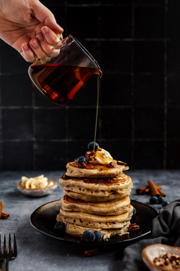 A person pouring syrup onto a stack of vegan banana pancakes with blueberries and a pat of butter on top, presented on a dark plate with fruit and spices scattered around.