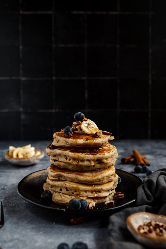 A stack of vegan banana pancakes with blueberries and banana slices on a dark plate with syrup drizzle, set against a dark backdrop with a rustic style presentation.