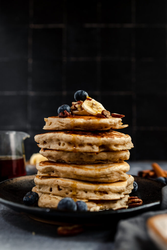 A stack of vegan banana pancakes with blueberries and syrup on a dark plate against a black tile background.