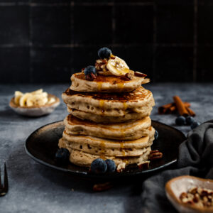 A stack of vegan banana pancakes with blueberries, banana slices, and syrup on a dark plate with breakfast ingredients in the background.
