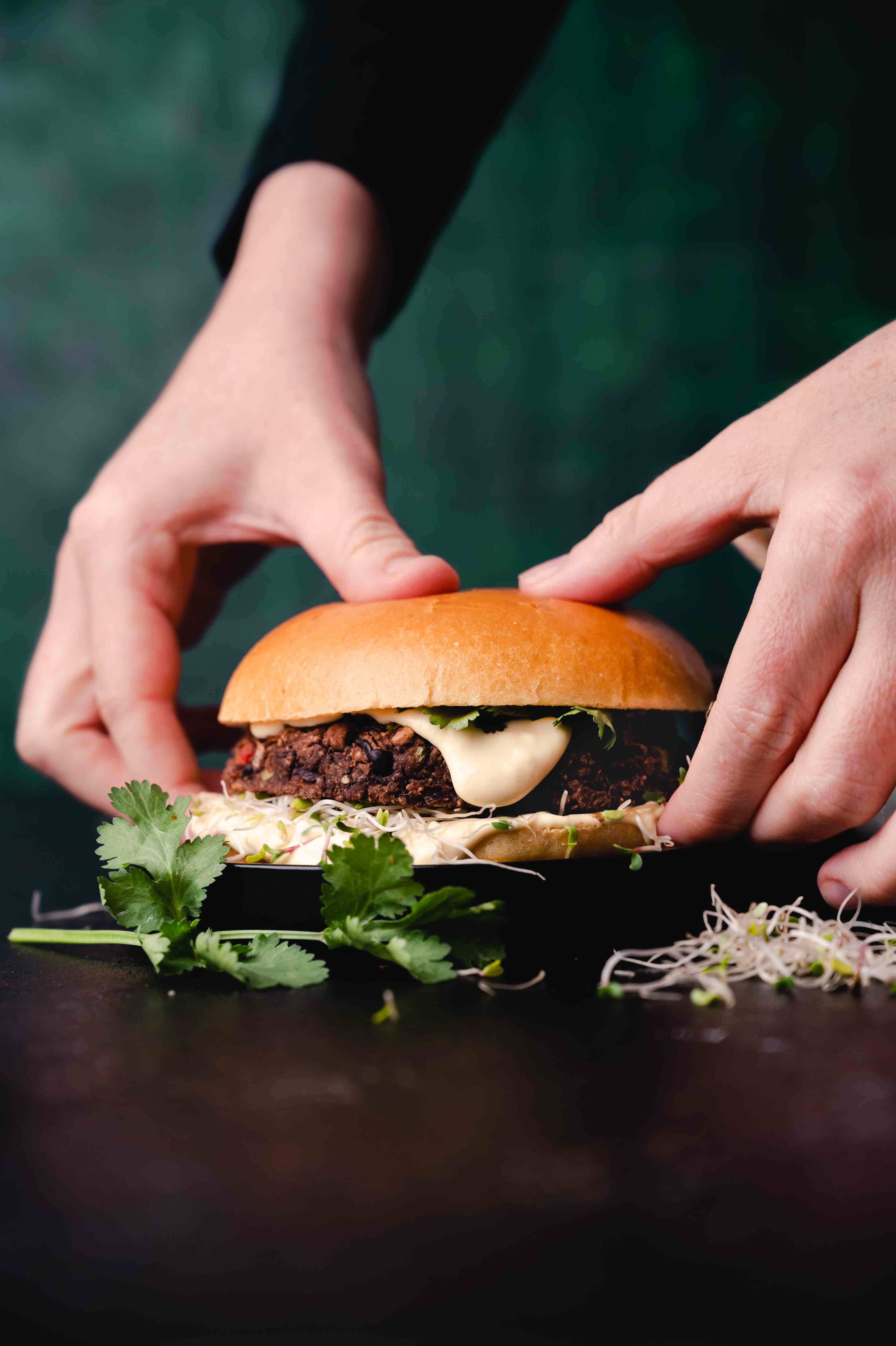 Hands presenting a gourmet burger with toppings and herbs on a dark background.