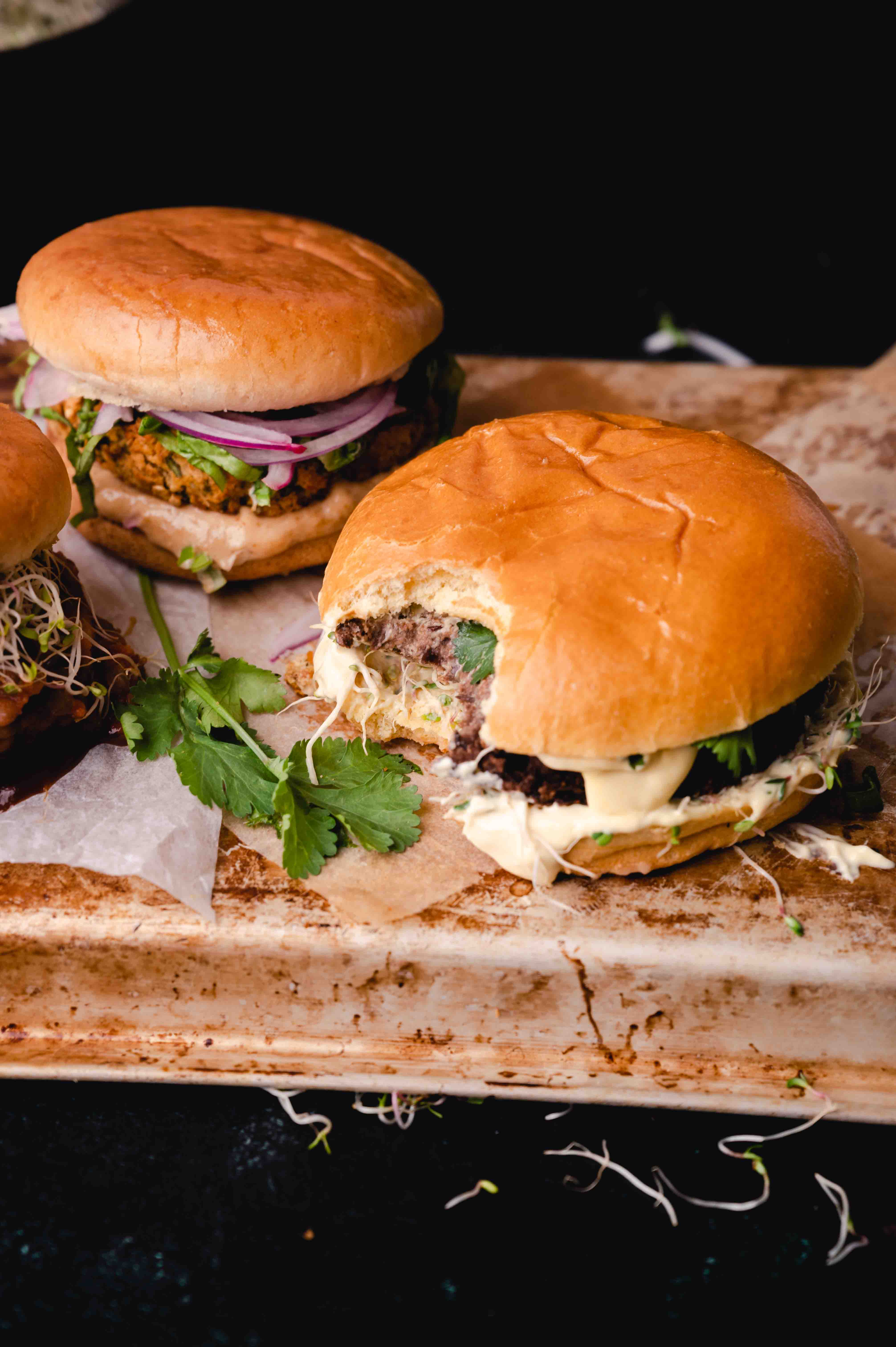 Two gourmet burgers with toppings on a rustic wooden board, accompanied by sprouts and herbs.
