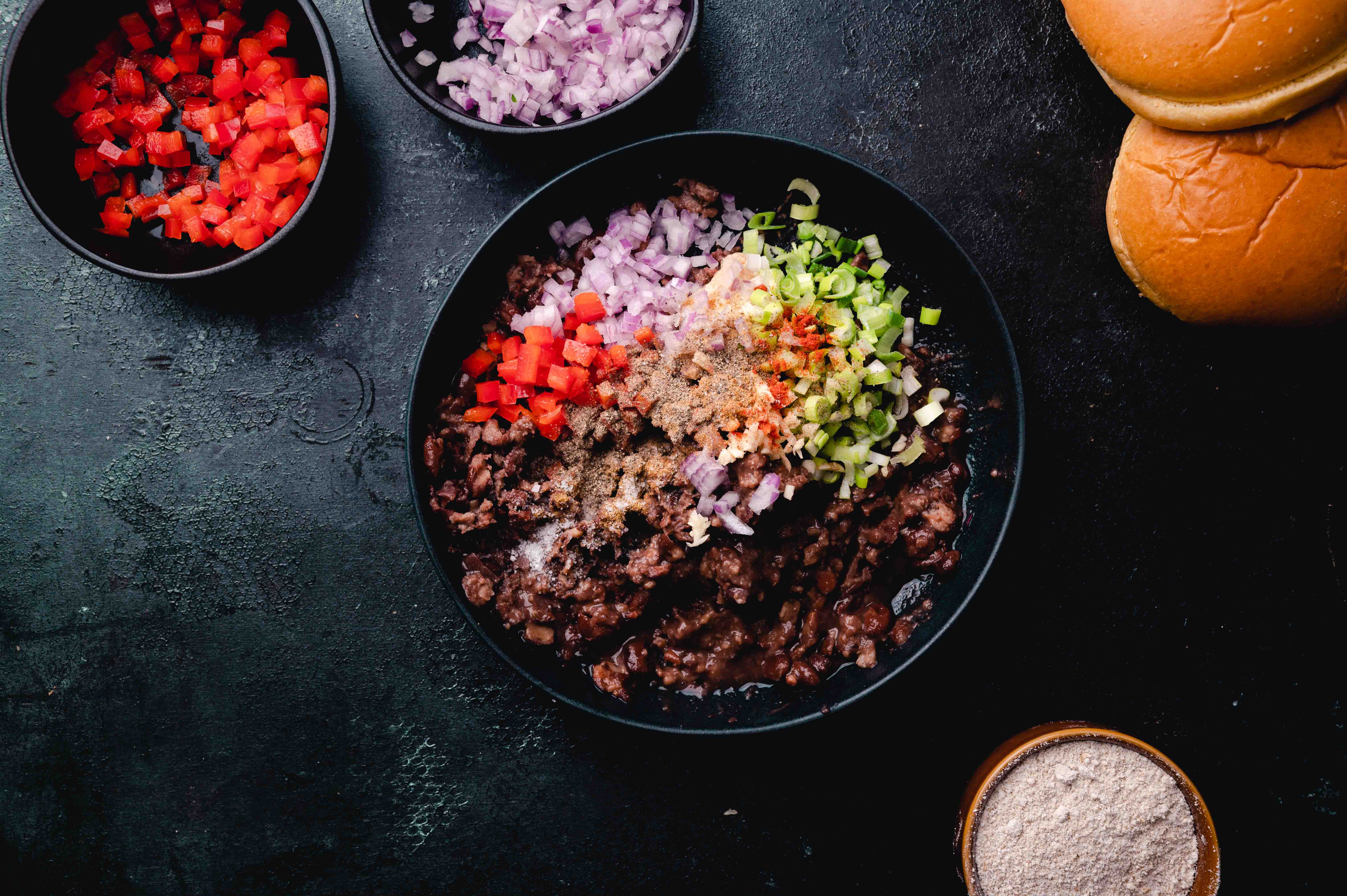 A black bowl filled with ground meat and diced vegetables on a dark table, surrounded by additional bowls of ingredients and burger buns.