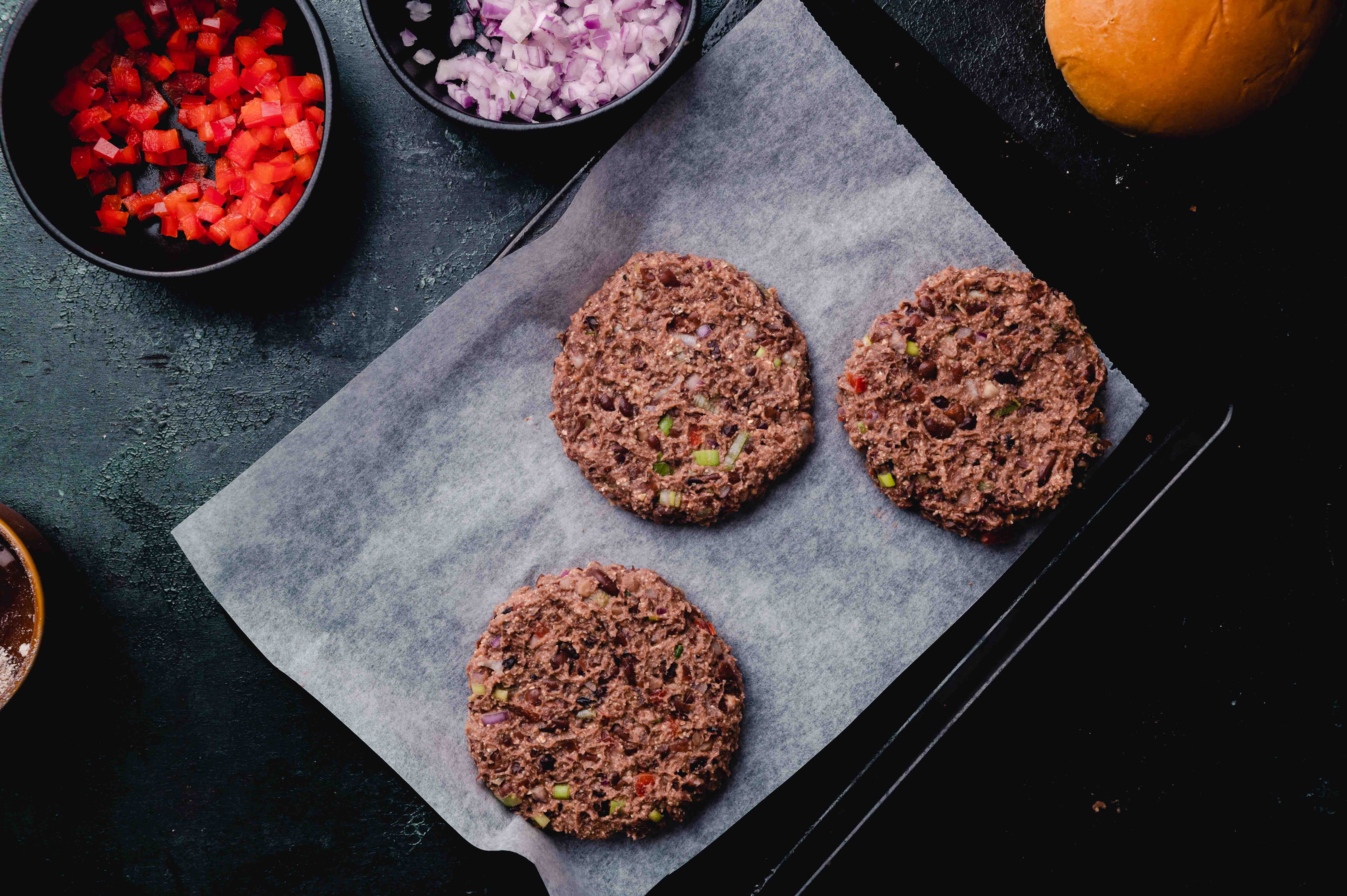 Three raw hamburger patties with diced vegetables on parchment paper, surrounded by chopped ingredients and a bun on a dark surface.