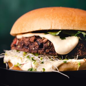 Close-up of a gourmet burger with a juicy patty, sprouts, herbs, and sauce on a black plate against a dark background.