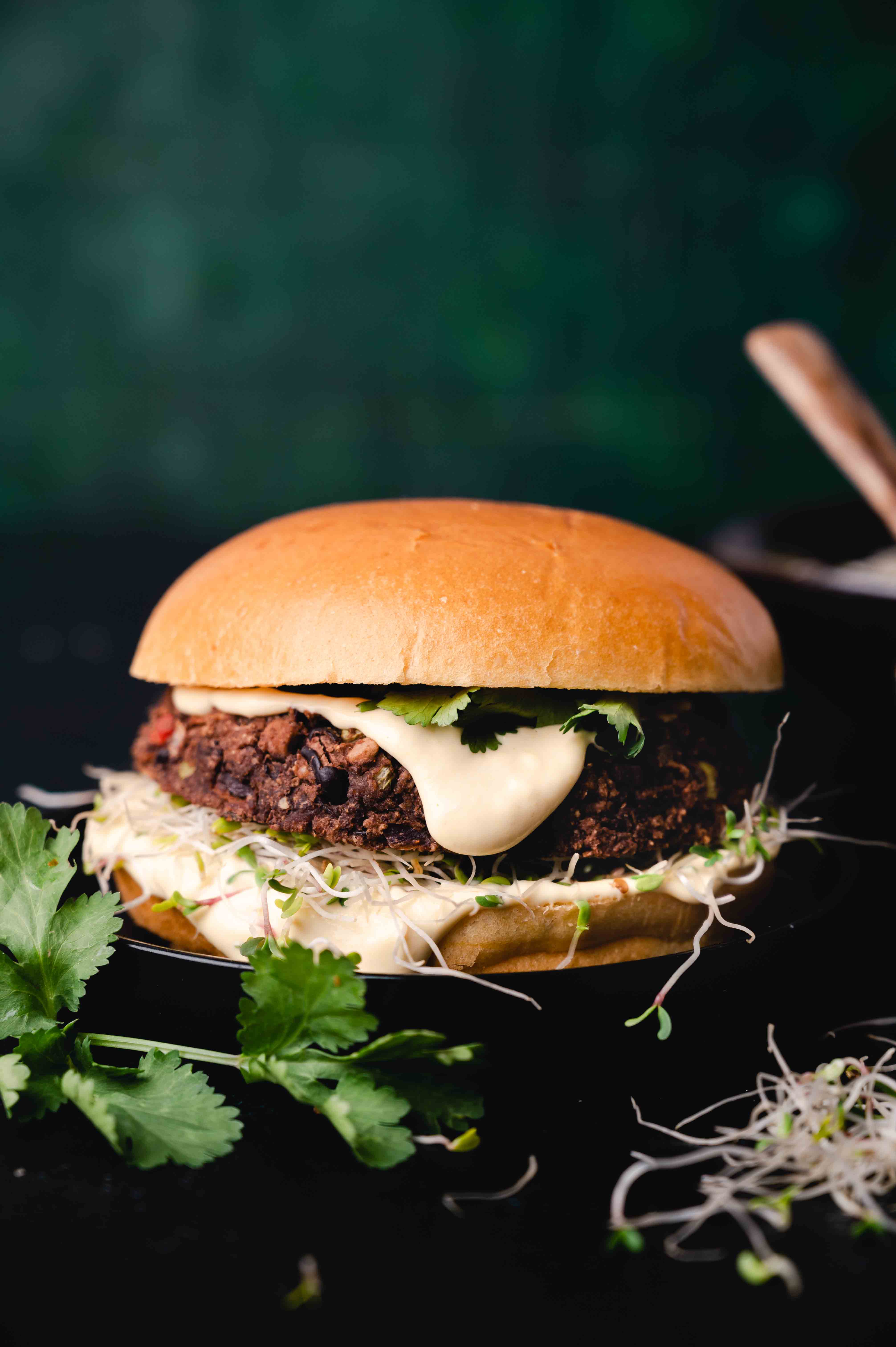 A veggie burger with a slice of cheese, lettuce, and sprouts on a black plate against a dark green background.